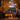 A two-story house is decorated for Halloween and surrounded by autumn foliage. The house is grey with dark shutters and a front porch. Jack-o'-lanterns are placed along the walkway leading to the front door, and lights glow warmly from the windows, creating a welcoming and festive scene. The lawn is covered with fallen autumn leaves.