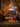 A two-story house is decorated for Halloween and surrounded by autumn foliage. The house is grey with dark shutters and a front porch. Jack-o'-lanterns are placed along the walkway leading to the front door, and lights glow warmly from the windows, creating a welcoming and festive scene. The lawn is covered with fallen autumn leaves.