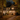A two-story house is shown at twilight, decorated for Halloween. The house has gray siding and a dark roof, and the yard is covered in fallen yellow and orange leaves. Orange pumpkins are placed on the porch steps and around the front yard. Lights are on inside the house and on the porch, creating a warm, inviting glow.