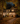 A two-story house is shown at twilight, decorated for Halloween. The house has gray siding and a dark roof, and the yard is covered in fallen yellow and orange leaves. Orange pumpkins are placed on the porch steps and around the front yard. Lights are on inside the house and on the porch, creating a warm, inviting glow.