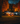 A one-story white house is decorated for Halloween at twilight. Orange, glowing pumpkins line the walkway leading to the front door. The surrounding trees are filled with vibrant orange and red autumn leaves, and the sky is a deep blue.