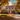 A two-story house is pictured in autumn with golden leaves covering the yard and driveway. The house is light-colored with a brown roof, shutters, and a decorated front door. Pumpkins and other fall decorations adorn the porch, adding to the cozy autumnal atmosphere.