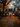 A two-story white house with black shutters is decorated for Halloween in autumn. Orange pumpkins and lanterns line a stone walkway leading to the front door. The yard and trees are covered in fallen leaves, with warm light emanating from the house windows, creating a cozy and festive scene.
