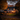 A two-story house is decorated for Halloween, framed by trees with orange and yellow leaves. Lit pumpkins are scattered across the front yard. Light shines from windows and porch lighting, creating a warm glow against the evening sky.