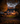 A two-story house is decorated for Halloween, framed by trees with orange and yellow leaves. Lit pumpkins are scattered across the front yard. Light shines from windows and porch lighting, creating a warm glow against the evening sky.