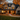 A two-story house is decorated for Halloween with pumpkins lining the walkway and porch. The house has a light beige facade with black shutters, a dark gray roof, and stone accents on the right side. Golden leaves fill the lawn and the trees surrounding the house, and warm light shines from the windows.