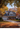 A two-story house is shown during the autumn season. The house is light-colored with a dark roof and black shutters, decorated for fall with pumpkins and gourds. The trees surrounding the house are full of colorful autumn leaves, creating a warm and inviting atmosphere.