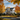 A two-story white house with black shutters is depicted in the fall. The house has a grey roof and is surrounded by trees with leaves in various shades of yellow, orange, and red. Pumpkins are visible on the front porch and in the garden, showcasing the autumn season.
