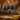 A gray two-story house is decorated for fall with pumpkins and a wreath on the front door. The lawn is covered in fallen autumn leaves. The trees surrounding the house are full of vibrant orange and yellow foliage, creating a warm and inviting atmosphere.