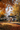 A white one-story house is framed by vibrant autumn foliage. Pumpkins and fall flowers decorate the porch and front yard. Fallen leaves cover the lawn and driveway in shades of orange and brown, creating a warm and inviting scene.
