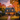 A two-story house is decorated for fall and Halloween with pumpkins lining the walkway and porch. The house is surrounded by trees with vibrant red, orange, and yellow leaves. Warm light shines from the windows, casting a golden glow on the scene beneath a twilight sky.