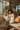 A smiling woman with curly hair sits cross-legged on a beige couch, bathed in sunlight. She wears white pajamas with gold trim and holds a delicate teacup. Behind her, a large window reveals a cityscape and an abundance of indoor plants, creating a warm and inviting atmosphere.