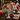 A warmly lit, elegantly set Christmas dining table fills the frame. A rustic wooden table is adorned with a green pine garland, bright red pillar candles in glass holders, and various red and silver Christmas ornaments, including a large patterned red bauble resting on a stack of grey-rimmed plates with polished silverware. In the softly blurred background, the twinkling lights of a Christmas tree add to the festive ambiance.