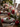 A beautifully set wooden dining table prepared for a holiday meal, viewed from a slight angle. The table is adorned with a festive evergreen garland, glowing red pillar candles, and various clear glass votives and lanterns, creating a warm, intimate atmosphere. Each place setting features a white textured dinner plate with a deep red napkin, silver cutlery, and a small pine sprig with frosted red berries and a silver bauble, while a blurred Christmas tree with warm lights is visible in the background.