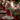 A beautifully decorated Christmas dining table features a rich red table runner, silver dinnerware with red charger plates, and both clear cut-glass and red stemmed drinking glasses. A lush green fir garland, adorned with red berries and ornaments, serves as a centerpiece, interspersed with glowing white pillar candles in clear glass hurricane holders. The background is softly blurred with warm, circular bokeh lights from a decorated Christmas tree, enhancing the festive ambiance.