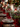 A beautifully decorated Christmas dining table features a rich red table runner, silver dinnerware with red charger plates, and both clear cut-glass and red stemmed drinking glasses. A lush green fir garland, adorned with red berries and ornaments, serves as a centerpiece, interspersed with glowing white pillar candles in clear glass hurricane holders. The background is softly blurred with warm, circular bokeh lights from a decorated Christmas tree, enhancing the festive ambiance.