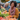 A smiling African-American woman stands at a farmer's market, holding a bunch of carrots. She is wearing an orange sweater and colorful scarf, surrounded by boxes of tomatoes, carrots, and leafy greens. Other shoppers and market stalls are visible in the background, creating a busy and colorful scene.