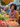 A smiling African-American woman stands at a farmer's market, holding a bunch of carrots. She is wearing an orange sweater and colorful scarf, surrounded by boxes of tomatoes, carrots, and leafy greens. Other shoppers and market stalls are visible in the background, creating a busy and colorful scene.