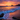 A breathtaking sunrise over a beach. The sky is a mix of orange and purple hues, illuminating the clouds. The waves are gently crashing onto the sandy beach, and dark rocks are visible in the foreground. The reflection of the sky paints the sand with a warm glow.