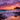 A vertical shot of a beach at sunset. Waves gently roll onto the sandy shore, lined with dark rocks. The sky is a vivid blend of orange, pink, and purple hues, with scattered clouds illuminated by the setting sun. A dark, mountainous landmass sits on the horizon under the vibrant sky.
