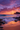 A vertical shot of a beach at sunset. Waves gently roll onto the sandy shore, lined with dark rocks. The sky is a vivid blend of orange, pink, and purple hues, with scattered clouds illuminated by the setting sun. A dark, mountainous landmass sits on the horizon under the vibrant sky.