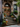 A brownstone entrance is decorated for autumn. A large wooden door with an arched top is adorned with two leafy wreaths. Stone steps lead up to the door, decorated with potted mums and small orange and white pumpkins. A stone handrail with plants cascading over it flanks the stairs, and a window above is decorated with greens.