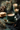 A close-up, warm-toned image features a dark teal ceramic mug filled with frothy coffee on a matching saucer, accompanied by a gold spoon. To the left, a stack of golden-brown cookies rests on a small silver plate. In the blurred background, lit candles in green holders, pine branches, and pine cones create a cozy, festive atmosphere, with scattered gold glitter on a dark, mottled surface.