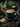 A close-up, slightly angled shot of a latte with detailed leaf art in a dark teal ceramic mug, resting on a matching saucer filled with scattered coffee beans. To the left, two chocolate chip cookies and a small golden spoon are arranged on a dark grey fabric. The background features blurred pine branches, pinecones, golden holiday ornaments, and a soft, glowing candle, creating a warm and festive atmosphere.