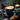A high-angle close-up shot of a dark teal ceramic mug filled with a coffee latte, featuring delicate white leaf-shaped latte art. The mug rests on a matching saucer with a small golden spoon, surrounded by dark brown cookies, scattered coffee beans, and brown sugar on a dark blue fabric. In the softly blurred background, evergreen pine branches, a lit candle, a pinecone, and a golden star ornament create a warm, festive, and inviting winter atmosphere.