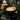 A close-up shot of a dark green ceramic cup containing a latte with intricate white latte art, possibly a rosetta pattern. The cup sits on a matching saucer on a dark fabric surface scattered with coffee beans and golden glitter, next to an elegant brass spoon. In the soft-focus background, a stack of chocolate chip cookies, a lit candle, and festive pine branches with bokeh lights are visible, creating a warm and cozy winter holiday atmosphere.