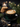 A close-up shot of a dark green ceramic cup containing a latte with intricate white latte art, possibly a rosetta pattern. The cup sits on a matching saucer on a dark fabric surface scattered with coffee beans and golden glitter, next to an elegant brass spoon. In the soft-focus background, a stack of chocolate chip cookies, a lit candle, and festive pine branches with bokeh lights are visible, creating a warm and cozy winter holiday atmosphere.