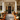 A beautifully decorated front porch greets visitors with autumnal charm. Pumpkins of various sizes line the steps, interspersed with planters overflowing with colorful mums. A black front door with a fall wreath is framed by white columns and a lantern, creating a warm and inviting scene.
