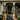 A grand Victorian house entrance decorated for autumn is shown. Pumpkins of various sizes and colors are arranged on and around the grey steps leading up to a dark, ornate front door. Planters filled with orange and white mums are placed on either side of the steps and on the porch. The facade of the house is a muted green, with intricate architectural details.