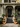 A grand Victorian house entrance decorated for autumn is shown. Pumpkins of various sizes and colors are arranged on and around the grey steps leading up to a dark, ornate front door. Planters filled with orange and white mums are placed on either side of the steps and on the porch. The facade of the house is a muted green, with intricate architectural details.