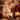 A warmly lit living room decorated for Christmas. In the foreground, a stone fireplace mantel is adorned with evergreen garland, glowing string lights, and five red and white knitted Christmas stockings hanging above a crackling fire visible through glass doors. To the left, a large, softly blurred Christmas tree twinkles with numerous warm lights and ornaments, surrounded by wrapped gifts, creating a cozy and inviting holiday atmosphere.