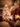 A warmly lit living room decorated for Christmas. In the foreground, a stone fireplace mantel is adorned with evergreen garland, glowing string lights, and five red and white knitted Christmas stockings hanging above a crackling fire visible through glass doors. To the left, a large, softly blurred Christmas tree twinkles with numerous warm lights and ornaments, surrounded by wrapped gifts, creating a cozy and inviting holiday atmosphere.