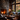 A cozy living room interior featuring a large stone fireplace with a bright fire. The room is furnished with comfortable armchairs and a sofa, and a large window offers a view of autumn leaves in warm hues. A wooden coffee table sits in the center of the room, adding to the rustic and inviting atmosphere.
