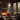 The interior of a rustic cabin is shown, featuring a stone fireplace with a burning fire, flanked by large windows showcasing colorful autumn trees. Dark brown leather furniture is arranged around a coffee table, and the room is lit by the fire and natural light filtering through the windows. The scene evokes a sense of warmth and tranquility.