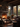 The interior of a rustic cabin is shown, featuring a stone fireplace with a burning fire, flanked by large windows showcasing colorful autumn trees. Dark brown leather furniture is arranged around a coffee table, and the room is lit by the fire and natural light filtering through the windows. The scene evokes a sense of warmth and tranquility.