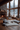 A beautifully decorated cozy living room in a rustic lodge, adorned for winter and the holidays. A large stone fireplace with a roaring fire warms the room, while a decorated Christmas tree and several lit lanterns with candles cast a soft, inviting glow. A plush white faux fur rug with multiple soft pillows and a tray holding two mugs and pastries creates a comfortable seating area on the wooden floor, all set against a stunning backdrop of a snow-covered evergreen forest visible through expansive windows.
