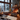 A warm and inviting rustic cabin interior during winter, showcasing a grand lit fireplace made of natural stone on the right and expansive windows on the left overlooking a snow-covered pine forest and distant mountains. The room is decorated with numerous lit candles in various lanterns, two small Christmas trees with string lights, and a comfortable seating area featuring cozy floor cushions and a white sheepskin rug on dark wooden floorboards, all bathed in a golden, ambient light.