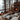 A warm and inviting rustic cabin living room is captured during winter, with large windows showcasing a snow-covered forest. A stone fireplace with a crackling fire and a decorated Christmas tree with warm lights are prominent features, surrounded by black lanterns holding lit pillar candles. Plush, patterned floor cushions and a white fluffy rug create a cozy seating area around a low wooden table set with a mug and snacks.