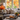 A living room interior featuring a gray sofa with decorative pillows. A coffee table in front of the sofa holds a tray with two pumpkins, two mugs and a vase with greenery and autumn leaves. A large window in the background showcases colorful autumn foliage.