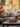 A living room interior featuring a gray sofa with decorative pillows. A coffee table in front of the sofa holds a tray with two pumpkins, two mugs and a vase with greenery and autumn leaves. A large window in the background showcases colorful autumn foliage.