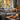 A cozy living room scene features a gray sofa with decorative pillows and a blanket. In front of the sofa, a wooden coffee table is decorated with small orange pumpkins, fall leaves, a mug, and lit candles. Through the window behind the sofa, colorful autumn foliage can be seen.
