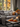 A cozy living room scene features a gray sofa with decorative pillows and a blanket. In front of the sofa, a wooden coffee table is decorated with small orange pumpkins, fall leaves, a mug, and lit candles. Through the window behind the sofa, colorful autumn foliage can be seen.