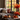 A cozy living room scene showcases autumn decor. A gray couch is adorned with textured pillows and a knitted throw. On a rustic coffee table, mini pumpkins, a lit candle, a mug, and decorative leaves are arranged. Behind the couch, a large window reveals the colorful foliage of autumn trees.