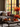 A cozy living room scene showcases autumn decor. A gray couch is adorned with textured pillows and a knitted throw. On a rustic coffee table, mini pumpkins, a lit candle, a mug, and decorative leaves are arranged. Behind the couch, a large window reveals the colorful foliage of autumn trees.