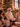 A heartwarming vertical shot of a senior man with a white beard, wearing a red Santa hat and a cream cable-knit sweater, laughing joyfully while holding a floral-patterned Christmas gift with a red ribbon. He is surrounded by blurred family members, including smiling children, and many festive presents scattered on the floor. In the background, a beautifully decorated Christmas tree with warm lights and ornaments adds to the cozy, holiday atmosphere, with a window visible on the right.