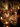 A multi-generational family gathered around a warmly lit dining table, enjoying a festive holiday dinner. A woman in a cream sweater smiles brightly, looking towards the right, while other family members, including an elderly man, laugh and converse. The table is adorned with plates of food, wine glasses filled with red wine, and glowing candles, all contributing to a cozy and joyful atmosphere with blurry golden string lights in the background.